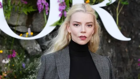 Anya Taylor-Joy attends the Dior Cruise 2025 show at Drummond Castle, Perthshire. She poses in front of a flower wall in a black top and a shirt with blonde hair and gold earrings.