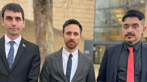 Emma Baugh/BBC Three men lined up outside court, all wearing jackets shirts and ties. Daniel Kidby has short dark brown hair; Ben Newman has short lighter brown hair and a close cropped beard; Beau Sebastien has a moustache and goatee.