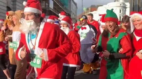 A closeup of people wearing red Santa outfits, with an elf, a penguin and a reindeer also in the crowd of participants in the Santa Dash.