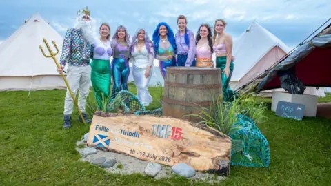 Tiree Music Festival A group dressed in sea-themed outfits and fantasy costumes pose beside a festival sign, barrel and fishing nets in front of large camping tents.