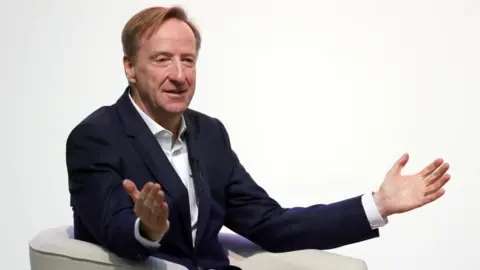 Reuters An older man with light, brown hair, dressed in a dark suit and white shirt and gesturing with open arms while sitting in a white chair.