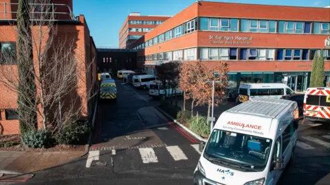 Several ambulances are parked outside St George’s Hospital in Tooting, South London. The large red-brick building has multiple windows and a sign reading ‘Grosvenor Wing Main Entrance’ above the entrance.