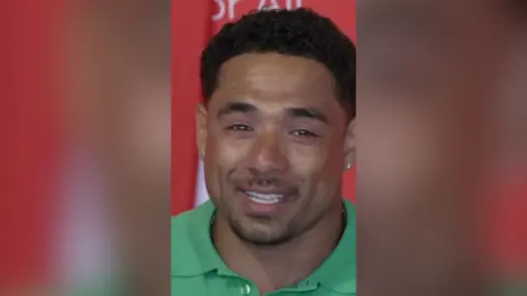 A man in a light green polo shirt is smiling while looking at the camera. There are tears in his eyes. The background is a red wall with unintelligible writing on it.
