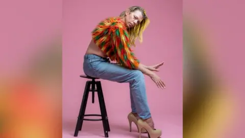 Tom Marshman wearing a multi colour feather half-jumper with denim jeans and cream stiletto heels. He is sat on a brown stool with his hands out in front, looking at the camera with a pink studio backdrop.