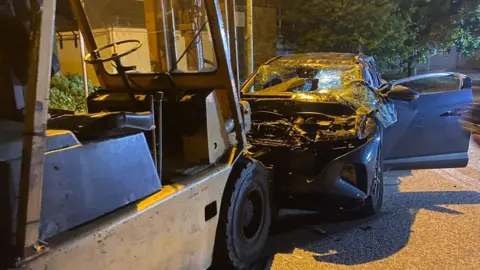 Newton Abbot Fire Station A badly smashed up car with an open passenger door, missing bonnet and wrecked windscreen sits in front of a forklift truck. It is night but the scene is lit with street lamps.