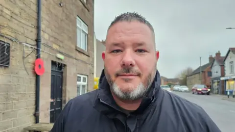 Keith Buzzard wears a black coat and  looks directly at the camera. To his left is a poppy on a drainpipe of a building. 