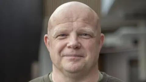 Andrew Bartlett Andrew Bartlett in the centre of the photograph smiling at the camera. He is middle aged, bald and has blue eyes. He wears a khaki long-sleeve t-shirt. 