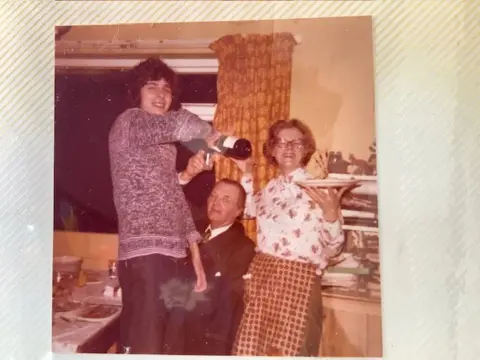 Cancer Research UK A faded photo in a scrapbook. There are brown/orange curtains behind in a classic 1970s kitchen with Peter on the left pouring himself a glass of wine, his father sitting down in the middle and his mum smiling and holding a plate up. All are in party mood