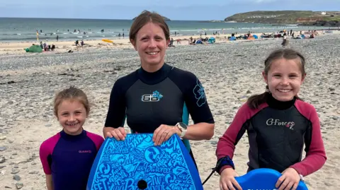 Mike Green Collette stands on a beach wearing a wetsuit and holding a body board with her two daughters who are doing the same. People on the beach can be seen behind them. 