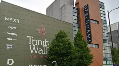 Shopping centre with large green building on the left of the image with the words Trinity Walk Shopping written on it and a large sign reading entrance next to a brick building with a long sign in black reading Debenhams