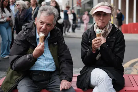 Chris Foote/BBC Scotland A middle-aged man and a woman sit on a street bench enjoying a couple of ice cream cones as the crowds pass by. The man wears a rain jacket and blue shirt as he takes a big lick of his cone, the woman, wearing a pink visor cap holds her mint choc chip cone.