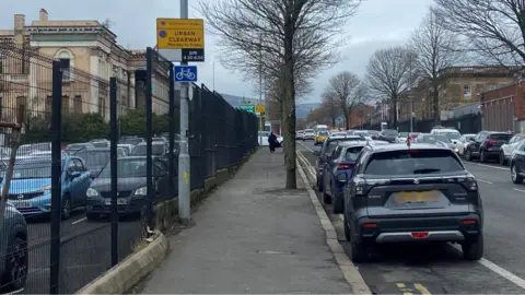 Three cars parked along a street in Belfast. A double yellow lane is on the left and a solid white line is on the right. A lamppost on the footpath has an urban clearway sign on it with a cycle sign below it.