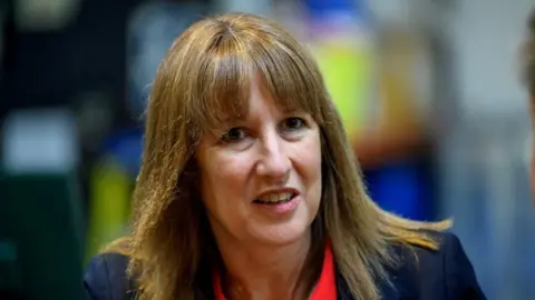 Reuters Rachel Reeves looks at the camera, with blue suit jacket and a red collared shift. 