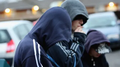 Generic close up image of three young people in hooded tops so their faces can't be seen. They are facing to the right and the middle one is smoking