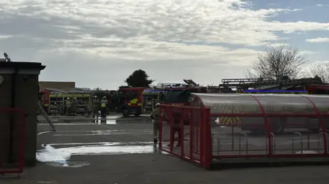 Ben Parker/BBC Multiple fire engines parked in a school playground. Water from hoses can be seen over parts of the concrete playground. Some firefighters walk around the area in their uniform.