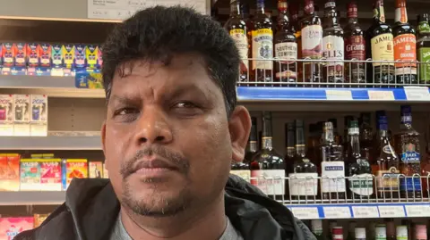 Man looking into the camera in a shop, with colourful bottles on display behind him. He has swept-back black hair and a neat moustache and beard.