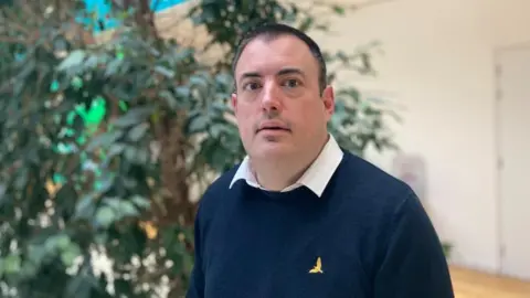 Laura Coffey/BBC Mike Hallam with short dark hair staring at the camera. He is wearing a blue sweater with a bird logo and a white shirt. He is in front of a large plant and a white wall.