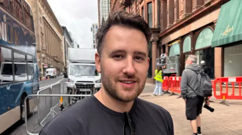 Matthew - a man with short dark hair and a thin beard, standing smiling on a city street. 