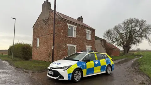 A marked police car parked on a country lane in front of a redbrick house in the countryside. The sky is overcast.