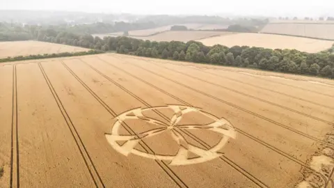 Lewis Johnstone Photography An aerial view of a crop circle resembling a five-spoked wheel with a rural landscape in the background