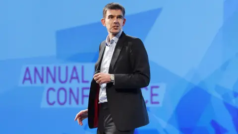 EPA Matt Brittin looks at the audience while standing and speaking onstage against a blue background with the words 'annual conference' on it, in London in 2016.