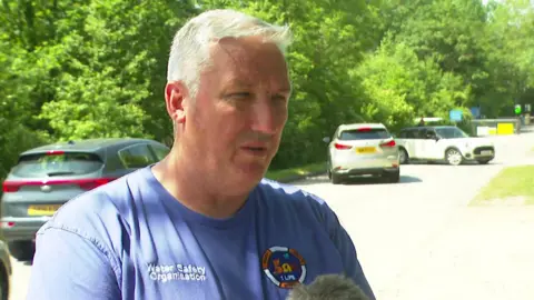 A middle-aged man with short white hair stands speaking into a media microphone. He wears a blue t-shirt with the words 'water safety organisation' on it. 