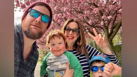 Dan, who is wearing sunglasses, poses for a photo with his wife, who is also wearing sunglasses, and their two children.