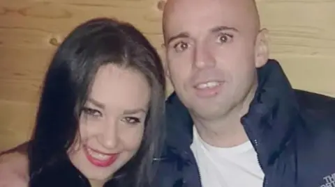 A male with a bald head wearing a white shirt and black puffer jacket, is pictured beside a woman with black hair. Behind them is a wood panelled wall. 