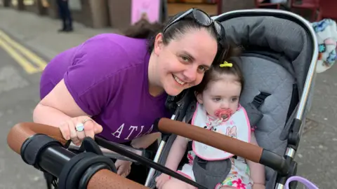 A woman is leaning over a baby in a buggy. She has brown hair pulled back and sun glasses perched on top of her head. She is wearing a purple t-shirt and smiling at the camera. The baby in the buggy has a yellow clip in her hair and a pink dummy in her mouth and is looking away from the camera.