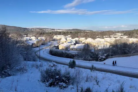 A snowy scene in Inverness. The picture has been taken looking down on a road. There are houses, their roofs covered in snow and two people are walking along a pavement with a dog walking behind them.