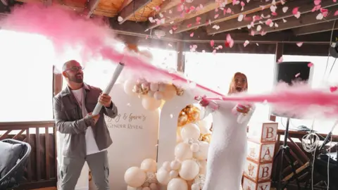 Photography by Charlotte Hayley and her partner are each holing long white tubes with pink powder coming out. They are laughing, Hayley is dressed in all white. Her partner has very short hair and a beard and wears glasses. He is wearing grey trousers and a jacket over a white t-shirt, 