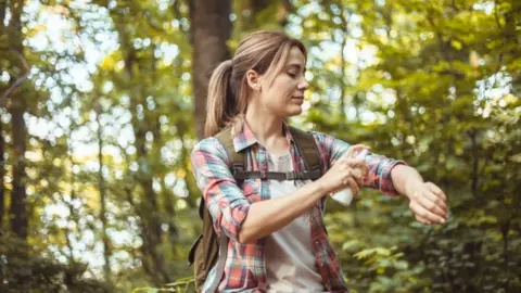 A woman with tied-back blonde hair sprays insect-repellent onto her left arm.