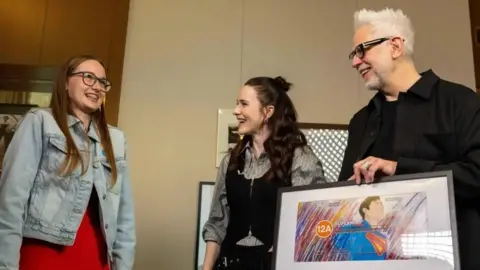 A girl in a denim jacket witha big smile as she talks to actor Rachel Brosnahan and director James Gunn in a room with soft, cream walls. James Gunn is holding a framed artwork depicting Superman in his blue superhero costume and an orange 12A age rating circle.