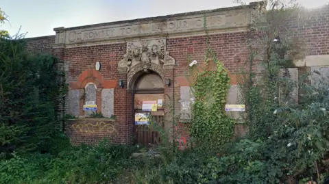 The former Romsey Labour Club is a one-storey brick building. The Google Streetview image shows its windows boarded up and trees and hedges growing in front of it.