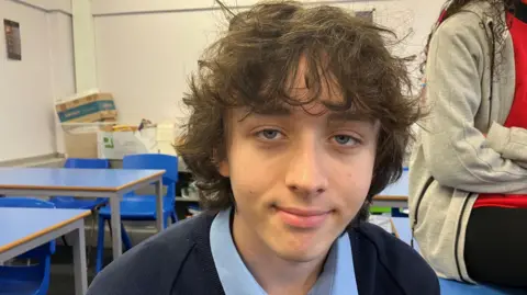 A teenage boy in a navy and blue school uniform, sitting in a classroom with other young people. He has brown wavy hair and blue eyes.