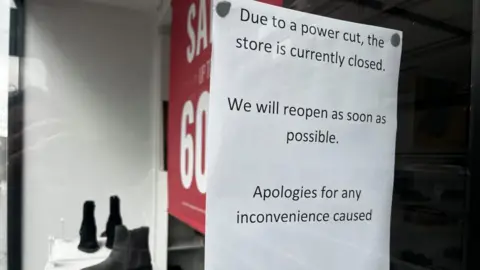 Alex Pope/BBC A sign in Clarks window, with a sale sign to the left and three boots. The sign is stuck on glass and is written in black on white paper. 