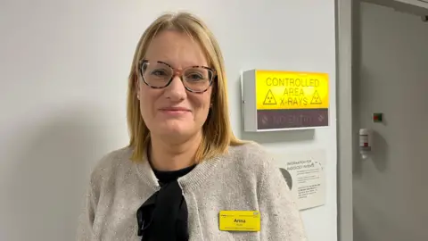 Ellen Knight/BBC Martin is photographed looking into the camera and smiling slightly. She is wearing a black blouse with a sparkly grey cardigan over the top. Martin has shoulder-length blonde hair, and is wearing a pair of tortoiseshell-framed glasses. She has a yellow NHS name badge pinned to her left lapel, which reads 'Anna' in black letters. Behind her left shoulder is a yellow, lit-up sign, which reads 'Controlled Area, X-Rays' in black letters. The walls of the room are white. 