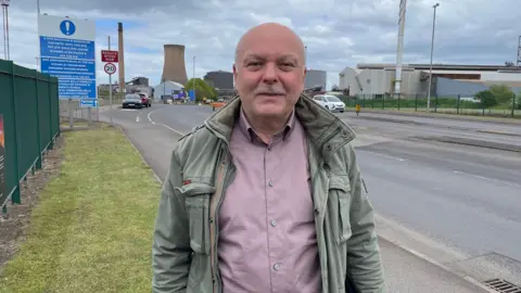A man with a bald head and grey beard stands in front of an industrial plant. He wears a mauve shirt and pale green jacket.