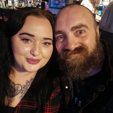 Llinos Hughes Llinos Hughes and her partner Paul smile at the camera. She has long dark hair and pink lipstick on, and smiles for the camera. Paul is also smiling, he has a shaved head and a dark brown beard.