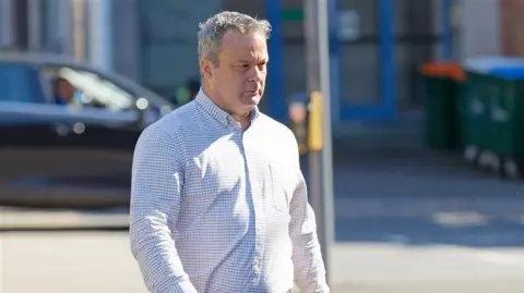 Christopher Elias walking in the sun to the court, he is wearing a white and grey checked shirt, has grey hair and has a furrowed brow. 