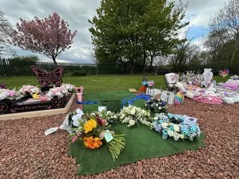 A collection of bouquets of flowers laid on a green mat beside the grave, which is open. Either side are colourful graves for other children, with trees including a pink cherry blossom in full bloom beyond.