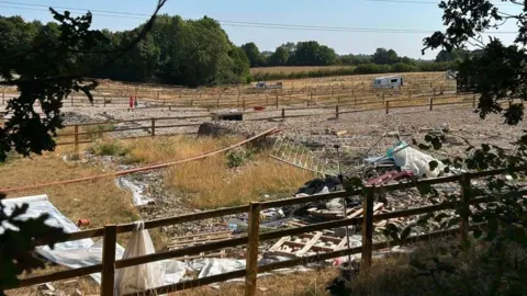 Supplied Abandoned land with hard standings and green/yellow grass growing. There is an abandoned rotating-style clothes line and rubbish in the foreground, and a caravan in the mid-distance. A lot of wooden three-bar fencing has been erected. There are green trees in the background.