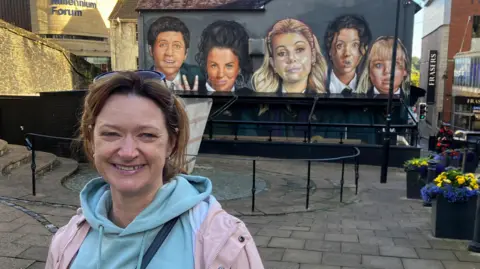 Julie Sargisson em frente ao mural Derry Girls, pintado na parede do frontão de um pub em Derry.