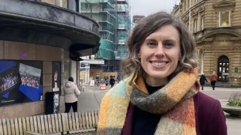 Jessica Toale has medium length brown hair and a multi-coloured scarf above a purple coat. She is stood in Bournemouth town centre.