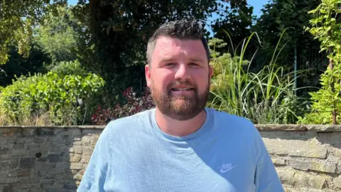 Ellen Knight/BBC Dickin photographed looking into the camera and smiling slightly. He is wearing a blue t-shirt and is stood in front of a stone wall which has plants and flowers growing along the top of it. There is strong sunshine and the sky is blue. 