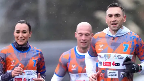 Danny Lawson/PA Three people running in a line. They have matching blue and orange sports tops on with "7" written all over them. Ennis-Hill on the left has black hair in a ponytail. Sinfield on the right is much taller, and his smiling at something off-camera. In the centre is a bald man.