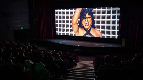 A cinema showing a film featuring a close up shot of a woman dancing with a full audience