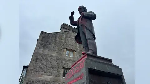 A side view of the red graffiti with the words 'coloniser' painted on the David Lloyd George statue. There is a building behind the statue with two windows. 