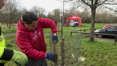 Ryan Simpson wears a red hoodie with the Freely Fruity logo in white across the front. He is helping to plant a tree