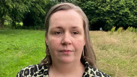 Julie McCloskey is standing in a large field with a number of purple plants and trees. She is wearing a leopard print top and has short brown hair.